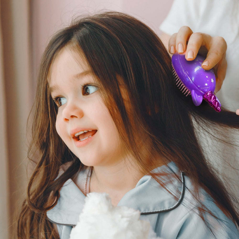 Smiling little girl having long hair brushed with a purple unicorn detangling brush for kids